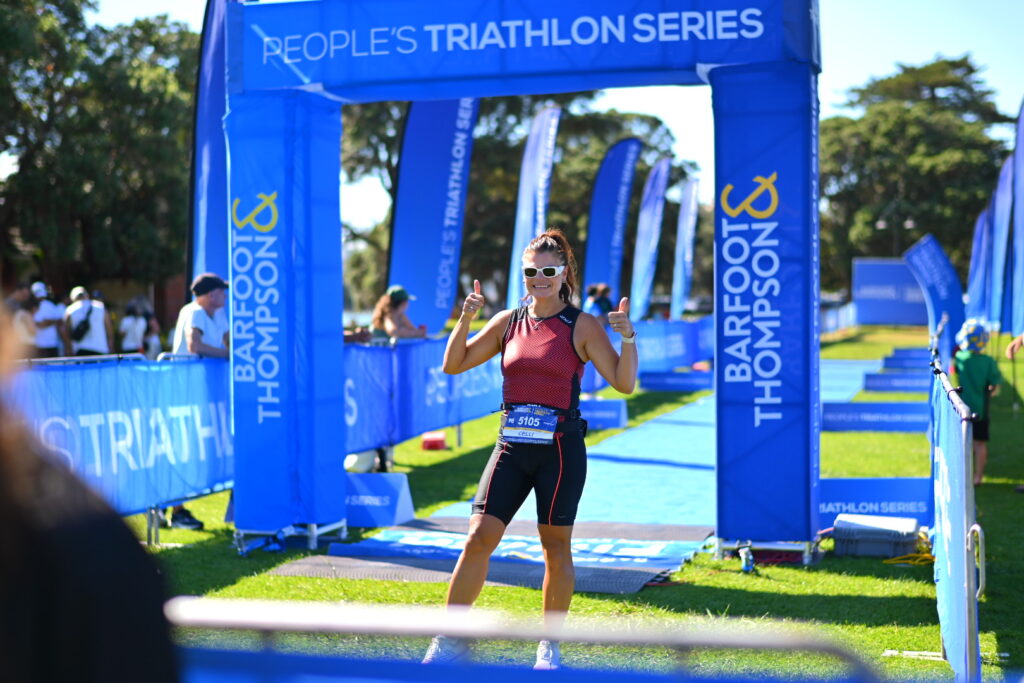 A smiling athlete celebrates at the blue People's Triathlon finish arch at Mission Bay.